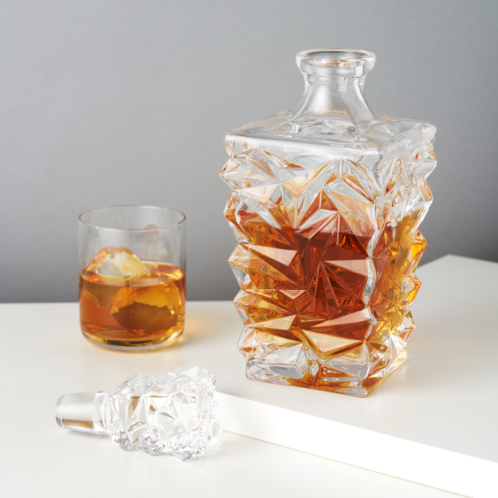 Decorative glass decanter with intricate design, filled with amber liquid, next to a glass with ice cubes on a light surface.