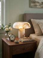 A mushroom-shaped table lamp made of polished white and beige marble sits on a dark wood bedside table next to a bed. The table also holds a small vase of white roses, a vintage-style alarm clock, a stack of books, and eyeglasses.