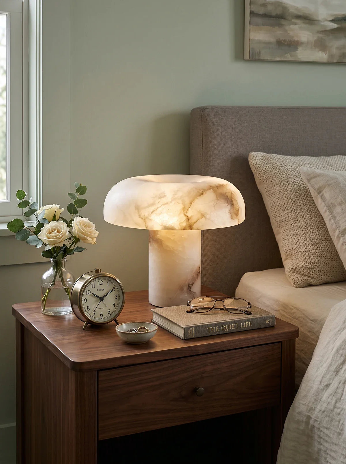A mushroom-shaped table lamp made of polished white and beige marble sits on a dark wood bedside table next to a bed. The table also holds a small vase of white roses, a vintage-style alarm clock, a stack of books, and eyeglasses.