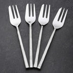 Four silver metal serving forks with long, textured handles arranged in a fan shape on a dark gray textured background.