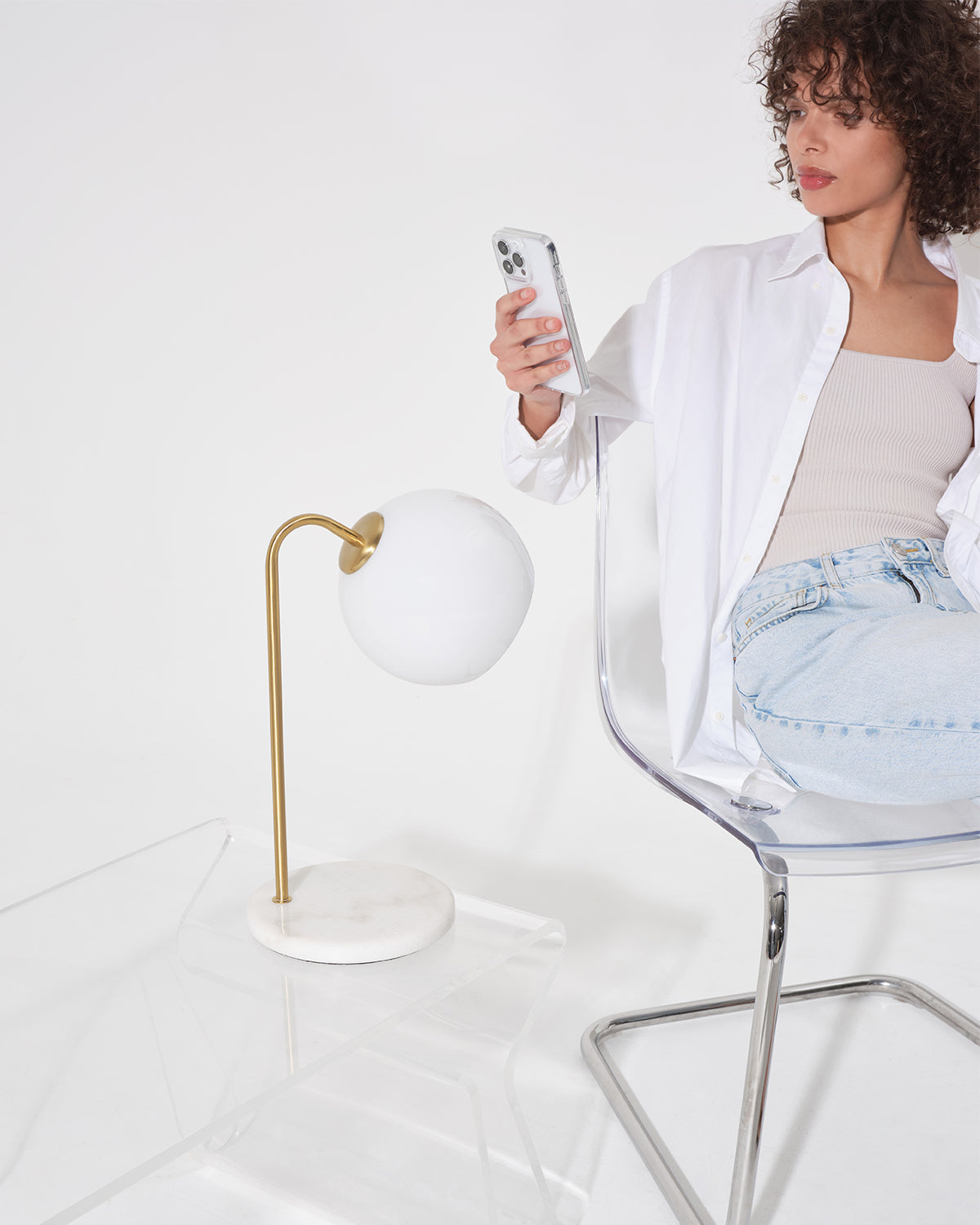 Woman sitting on a chair holding a phone next to a white table lamp with a gold base.