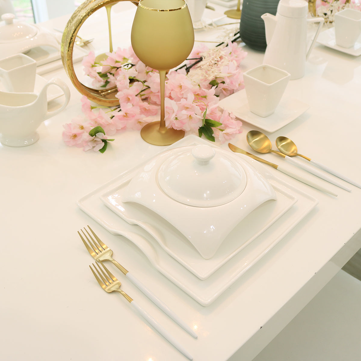 Modern table setting with white square dinnerware, gold cutlery, and pink floral centerpiece