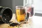 Glass teacup with tea and a container of loose tea leaves on a wooden surface.