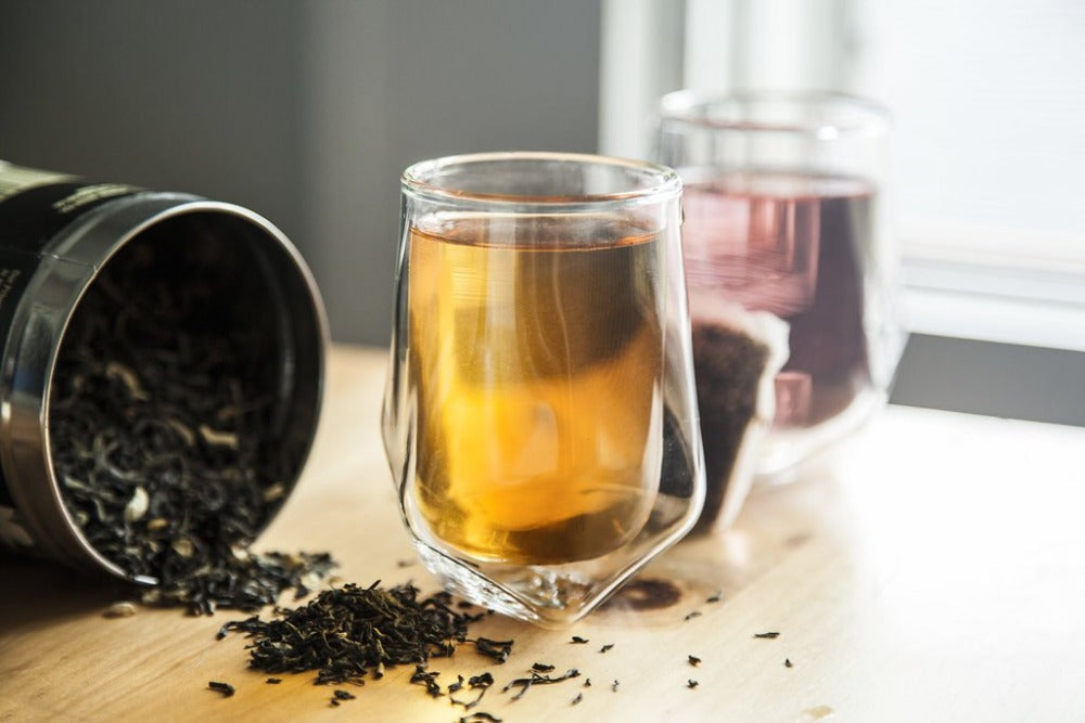 Glass teacup with tea and a container of loose tea leaves on a wooden surface.