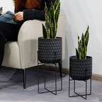 Two black textured planters with snake plants on a gray floor next to a person sitting on a white chair.