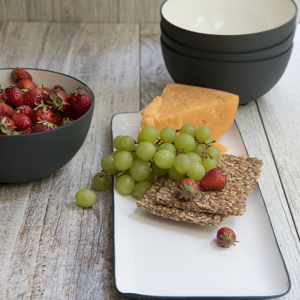 Platter with grapes, cheese, and crackers next to a bowl of strawberries on a wooden surface.