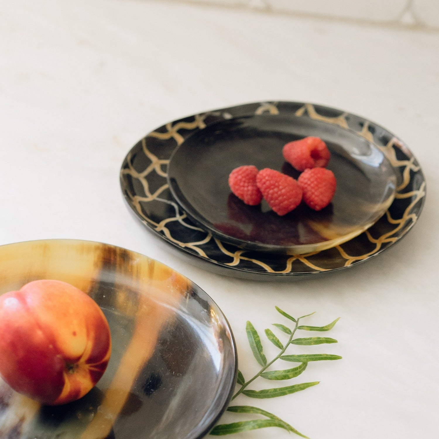 Two decorative plates with a black and gold pattern, one containing raspberries and the other an apple, on a white surface.