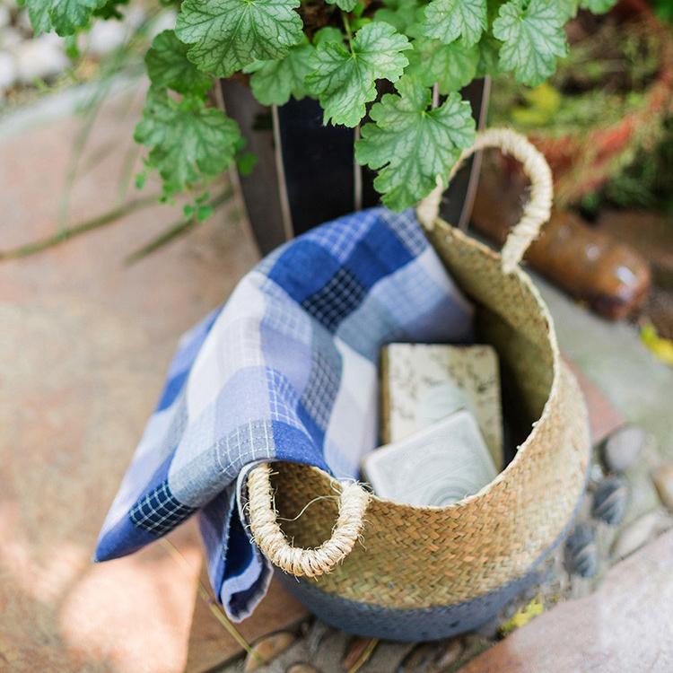 Woven basket with a blue checkered towel and soap inside, surrounded by plants.